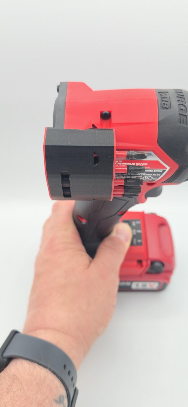 May include: Red and black power drill with a black attachment. The drill has the text "M18 SURGE" and "HYDRAULIC DRIVER" printed on it. The drill is held by a person.