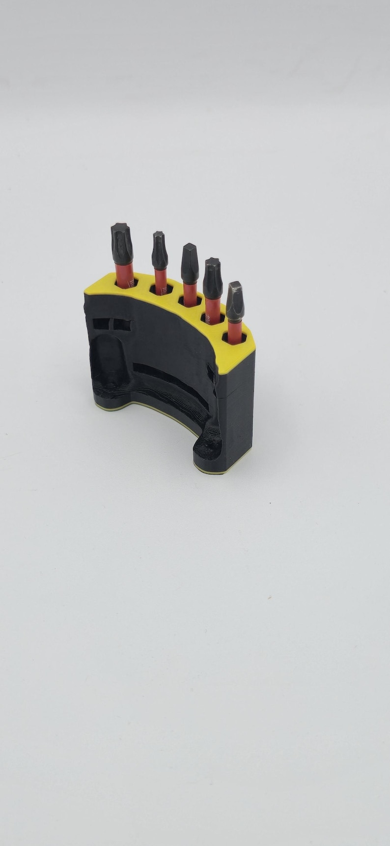 May include: A black and yellow bit holder with five dark metal bits. The holder is curved and has a yellow interior. The bits are arranged in a row, each with a red base.