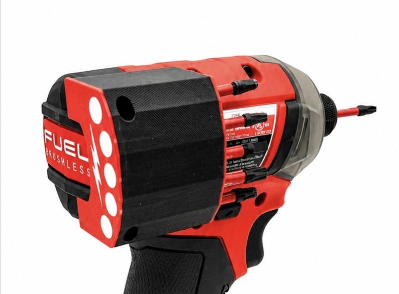 Puede incluir: Un atornillador de impacto sin escobillas FUEL rojo y negro. La herramienta tiene un mango negro y un cuerpo rojo con la palabra "FUEL" y un logotipo de rayo. La herramienta tiene una cubierta de pl&aacute;stico transparente y una broca negra.