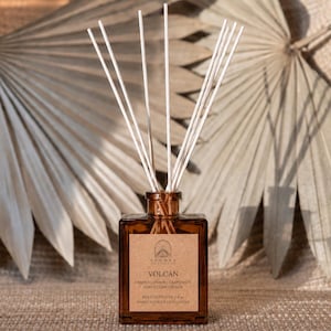 May include: A square amber glass reed diffuser bottle with a tan paper label reading VOLCÁN, ORANGE | LEMON | GRAPEFRUIT, AGAVE | LIME | PEACH, REED DIFFUSER 4 oz, HAND POURED IN ILLINOIS, featuring several thin white reeds extending from the top.