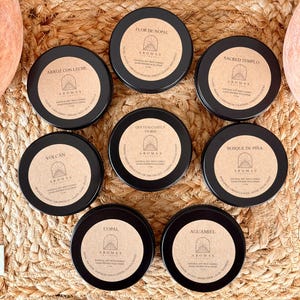 May include: A collection of eight black, round soy wax candles with brown labels. Each label features the word "AROMAS" and a unique scent name, including "Flor de Nopal" and "Arroz con Leche". The candles are arranged on a woven surface.