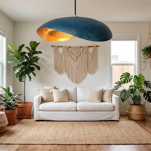 Könnte beinhalten: Ein modernes Wohnzimmer mit einer großen, blau-goldenen Pendelleuchte. Ein weißes Sofa mit beigen Kissen steht auf einem Jute-Teppich. Topfpflanzen und ein Makramee-Wandbehang ergänzen die Einrichtung.
