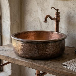 May include: A round, hammered copper vessel sink with a matching metallic tap. The sink features a textured, dimpled surface and a warm, reddish-brown finish. It is mounted on a wooden surface alongside a small soap dish and a folded cloth.