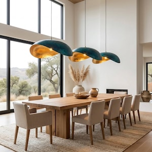 Puede incluir: Un comedor moderno con una gran mesa de madera y ocho sillas tapizadas en beige. Sobre la mesa cuelgan tres lámparas colgantes de color verde azulado y dorado. La habitación tiene grandes ventanales con vistas a un paisaje natural, con una alfombra neutra.