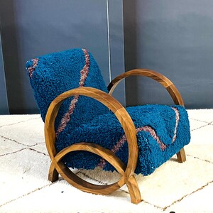 May include: A teal blue armchair with a curved wooden frame and a plush, textured fabric. The chair is sitting on a white rug with a diamond pattern.