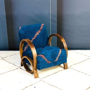 May include: A blue armchair with a wooden frame and a plush, blue and pink patterned cushion. The chair is sitting on a white rug with a geometric pattern.