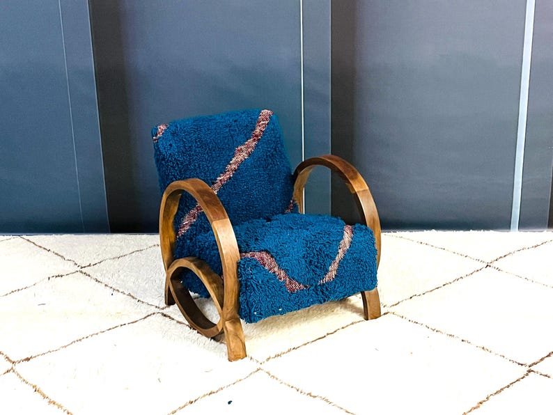May include: A blue armchair with a wooden frame and a plush, blue and pink patterned cushion. The chair is sitting on a white rug with a geometric pattern.