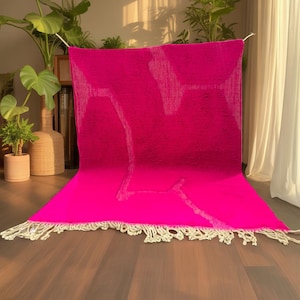 May include: A vibrant fuchsia rug with a textured surface and a cream-colored fringe. The rug has a geometric design in a lighter shade of pink. The rug is displayed on a wooden floor, with plants and curtains in the background.