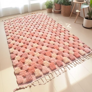May include: A rectangular rug with a textured, tufted design in shades of pink and cream. The rug features a grid-like pattern of raised, pom-pom-like elements. The edges are finished with a fringe detail. The rug is placed on a light-colored wooden floor.