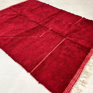Puede incluir: Una alfombra marroqu&iacute; de color rojo intenso con un patr&oacute;n geom&eacute;trico y flecos blancos. La alfombra est&aacute; hecha de lana y tiene una textura suave y mullida.