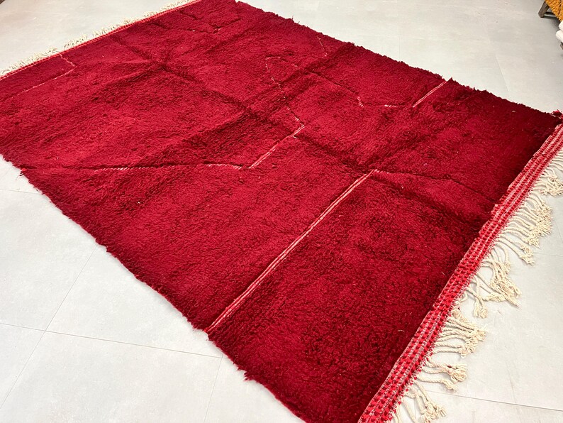 Puede incluir: Una alfombra marroqu&iacute; de color rojo intenso con un patr&oacute;n geom&eacute;trico y flecos blancos. La alfombra est&aacute; hecha de lana y tiene una textura suave y mullida.