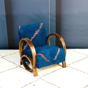 May include: A blue armchair with a wooden frame and a plush, blue and pink patterned cushion. The chair is sitting on a white rug with a geometric pattern.