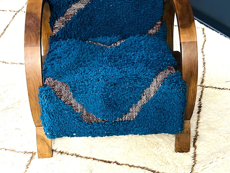 May include: A teal blue and burgundy patterned cushion sits on a wooden chair with curved arms. The chair is on a white and brown rug.