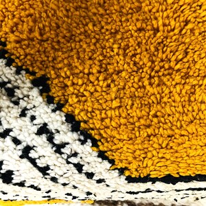 May include: Close-up of a yellow, white, and black woven rug with a geometric pattern. The rug is made of soft, fluffy fibers.