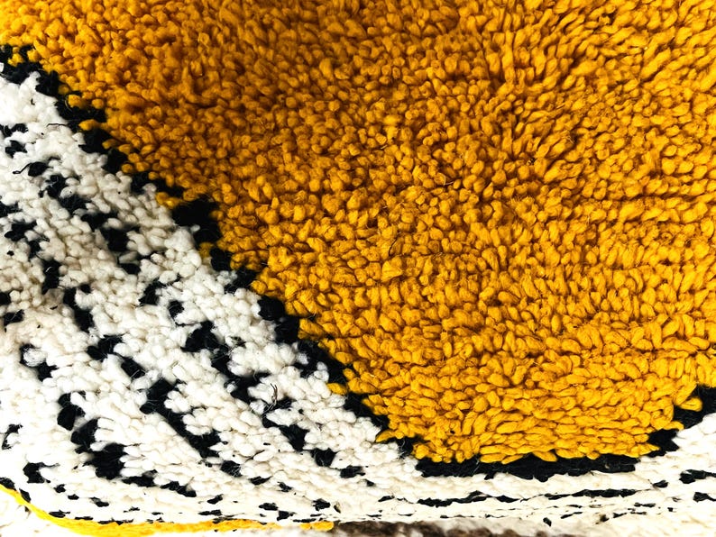 May include: Close-up of a yellow, white, and black woven rug with a geometric pattern. The rug is made of soft, fluffy fibers.