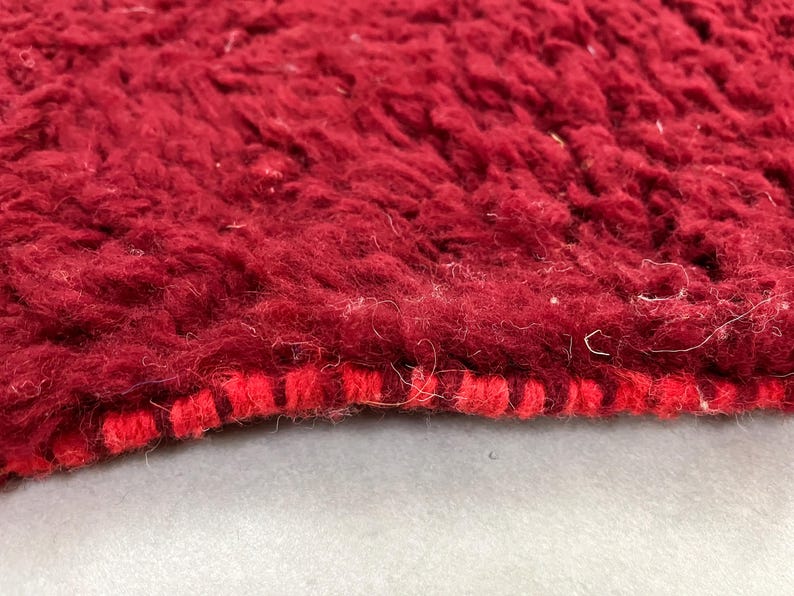 Puede incluir: Primer plano de una alfombra de color rojo intenso con un borde tejido rojo y blanco. La alfombra tiene una textura suave y peluda.