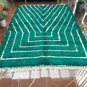 May include: A rectangular, emerald green rug with a geometric white pattern. The rug features a series of white, angled lines that create a chevron design. The rug has a fringe along the bottom edge. This rug is suitable for a living room or bedroom.