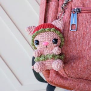 May include: A pink crocheted animal keychain with a green and red hat attached to a pink backpack.
