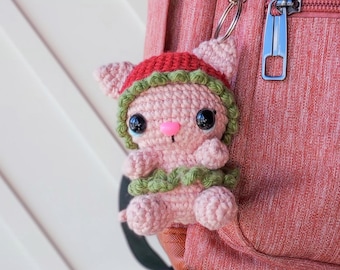 Pode incluir: Um chaveiro de animal de crochê rosa com um chapéu verde e vermelho preso a uma mochila rosa.