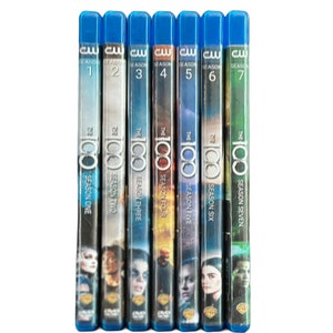 Puede incluir: Una colección de siete estuches de DVD Blu-ray de la serie de televisión "The 100". Cada estuche tiene una parte superior azul y muestra el número y el título de la temporada. Los estuches están dispuestos verticalmente, mostrando los lomos con el logotipo y las imágenes de los personajes.