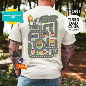 May include: Beige t-shirt with a colorful road map design on the back, featuring houses, roads, and a circular intersection. The shirt also has the text "TIRED DAD CLUB -[EST.2025]-." and "PERSONALIZE ME!"