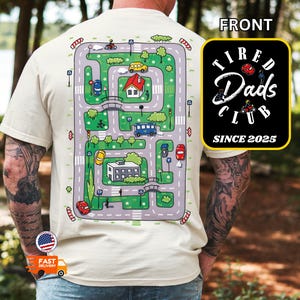 May include: Beige t-shirt with a colorful road map design on the back, featuring cars, buildings, and a bridge. The front has a black square with the text "Tired Dads Club SINCE 2025".
