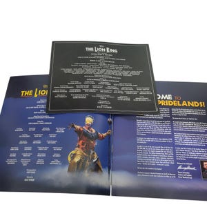 May include: Open book from the Lion King musical. The cover features the title and cast credits. Inside, a colorful image of a performer in costume is visible. The pages are blue and white with text.