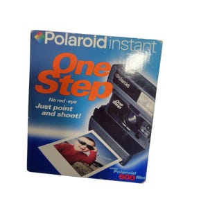 Peut inclure: Boîte d'appareil photo instantané Polaroid OneStep vintage. La boîte est bleue avec du texte orange qui dit "One Step" et "Just point and shoot!". Une photo d'un enfant portant des lunettes de soleil est sur le devant. La boîte utilise du film Polaroid 600.