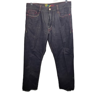 May include: Dark wash denim jeans with colorful stitching accents. The jeans feature red and yellow stitching on the pockets and seams, with the brand name "Coogi" embroidered on the waistband. The jeans are hanging on a black hanger.