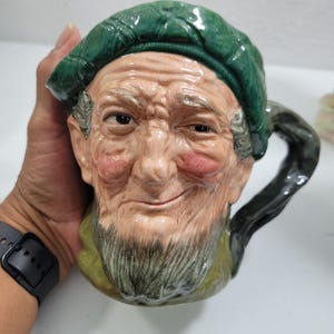 Op de afbeelding: Keramische mok in de vorm van het hoofd van een oude man, met een groene pet, grijze baard en roze wangen. De mok heeft een zwart handvat en wordt door een hand vastgehouden.