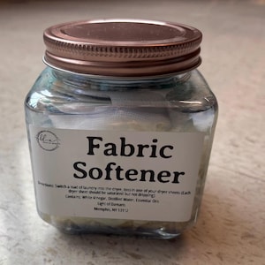 Peut inclure: Un bocal en verre transparent avec un couvercle en bronze, rempli de liquide et de feuilles de tissu blanches. L'étiquette indique "Adoucissant Textile". Produit de nettoyage ménager.