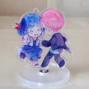 Puede incluir: Un soporte acrílico transparente con una ilustración de dibujos animados de dos figuras abrazándose. Una figura tiene el pelo azul y un vestido azul, la otra tiene el pelo rosa y una chaqueta morada. El soporte tiene una base redonda.