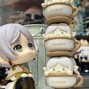 以下が含まれることがあります： アニメスタイルのフィギュアのコレクション。1つのフィギュアは座っており、3つのフィギュアがティーカップの中に積み重ねられています。ティーカップは白く、「Frieren」の文字がプリントされています。フィギュアは白い髪とエルフのような耳をしています。