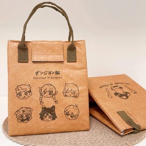 Könnte beinhalten: Braune Papiertüte mit Klappenverschluss und grünen Griffen. Die Tasche zeigt Illustrationen von Cartoon-Charakteren und japanischen Text, der "Delicious in Dungeon" lautet. Daneben liegt eine passende gefaltete Tasche.