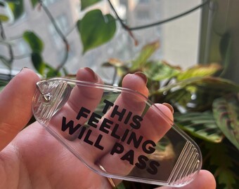 May include: A clear plastic keychain shaped like a hotel key with the text "This Feeling Will Pass" printed in black.