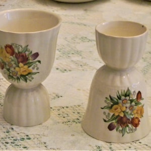 May include: Two white ceramic egg cups with floral designs. The egg cups have a fluted design and feature a bouquet of yellow, orange, and purple flowers with green leaves.
