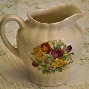 May include: A white ceramic creamer with a floral design featuring yellow, red, and purple tulips, green leaves, and white flowers. The creamer has a rounded body and a handle.