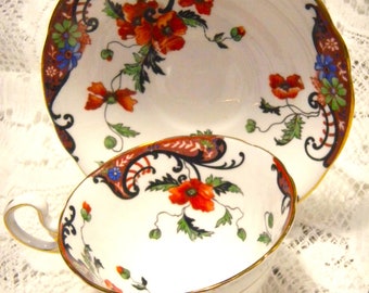 May include: A white teacup and saucer with a floral pattern. The pattern features red poppies, green leaves, and black and blue swirls. The edges of the cup and saucer are trimmed with gold.