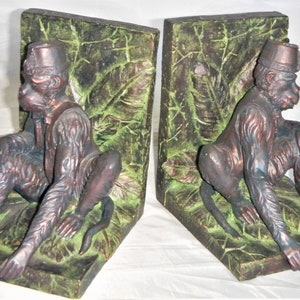 May include: A pair of bookends featuring a bronze monkey figurine with a red fez hat sitting on a green leafy base.