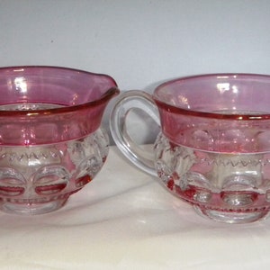 Puede incluir: Juego de dos piezas de fuentes de vidrio transparente con borde y asa rosas. La mitad inferior de las fuentes tiene un patrón decorativo. Una fuente tiene un pico, la otra no.