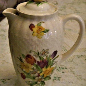May include: A white ceramic teapot with a floral design. The teapot has a lid with a yellow flower on top. The flowers on the teapot are yellow, purple, and red.