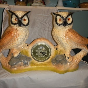 May include: A vintage owl-shaped clock with two ceramic owls perched on either side of a clock face. The owls have orange eyes, black accents, and tan and brown feathers. The clock face is green with black hands.