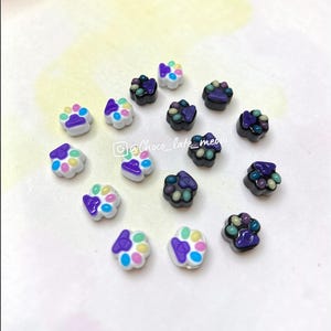 May include: A collection of small paw print-shaped charms in white and black bases with colorful accents. Each charm features a central pad and four toes in shades of blue, pink, yellow, and purple. The text @Choco_late_meow is visible.