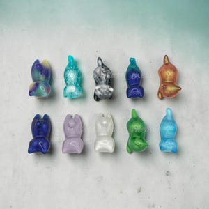 May include: Ten small, smooth, cat-shaped figurines arranged in two rows. The figures feature various colors and finishes, including marbled, translucent, and solid tones like blue, purple, white, black, green, and orange, all in a seated pose.