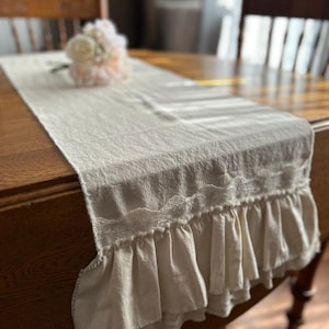 May include: A beige linen table runner with a lace trim and ruffled edges. The runner is on a wooden table with a bouquet of pink flowers.