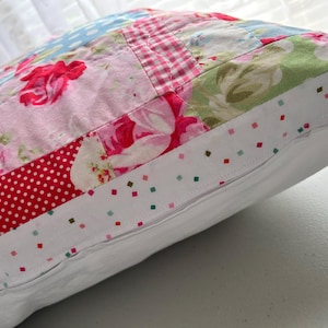 May include: A patchwork pillow with a floral pattern in pink, red, and green. The pillow has a white border with small, colorful square accents and a zipper closure. The pillow is a decorative home accent.