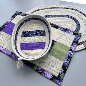 May include: A rectangular fabric placemat with purple floral patterns and text, a matching oval rope basket, and an oval rope mat. The fabric features text including A Time to Gather Stones, A Time to Build Up, A Time to Heal, and A Time to Dance.