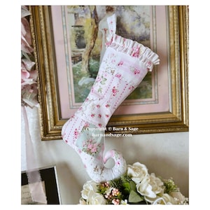 May include: A fabric Christmas stocking featuring a patchwork design of pink roses, green leaves, and white stripes. The stocking has a curled toe and a ruffled cuff at the top. The text "Copyright © Barn & Sage barnandsage.com" is visible.