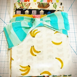 May include: A collection of fabric pieces with various patterns, including bananas, houses, and floral designs. A large bow with turquoise and white stripes is on top. The fabrics are arranged on a cutting mat.