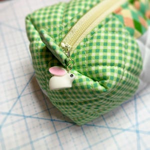May include: A green and white gingham quilted pencil case with a cream-colored zipper. A small white bunny charm with pink ears hangs from the zipper pull. The case is on a light blue grid surface.